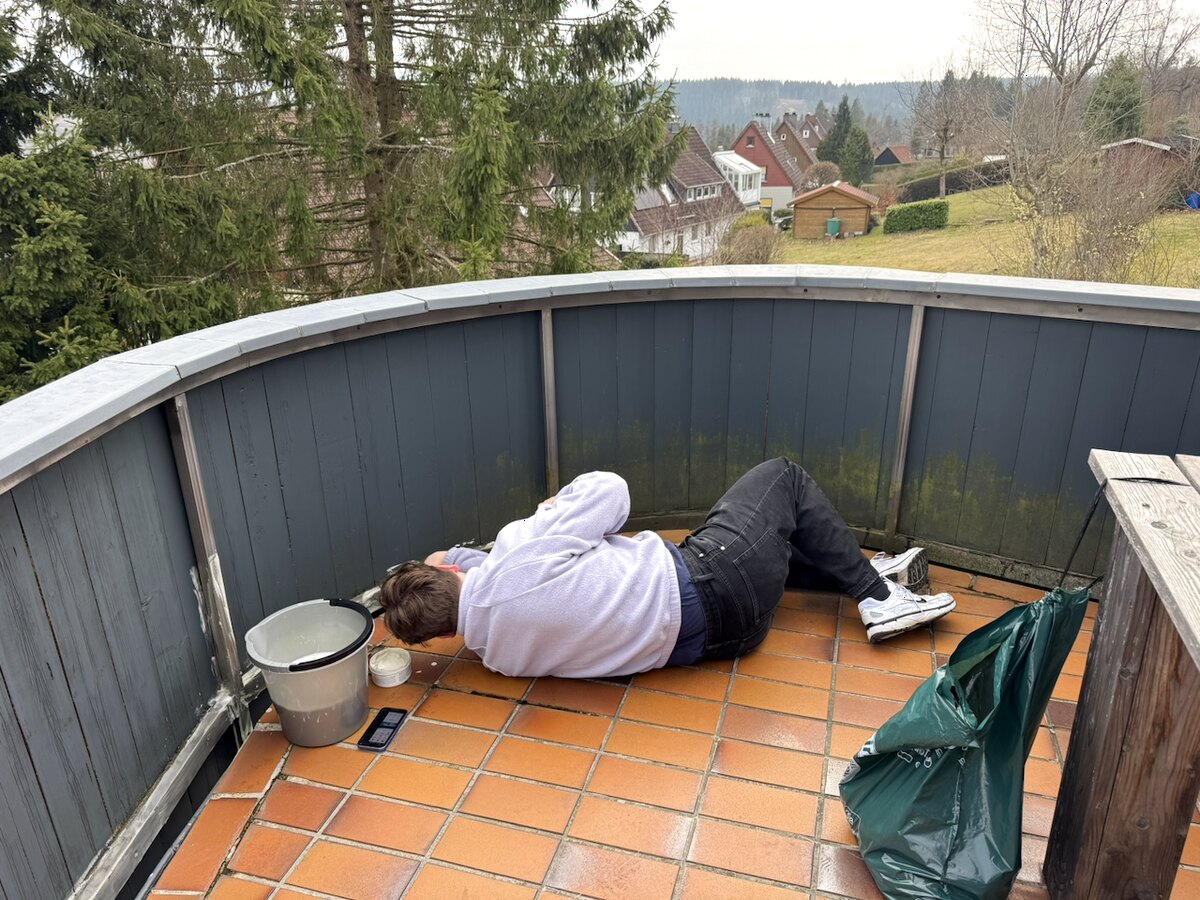 Reinigungskraft entfernt am Boden liegend den Schmutz am Sockel einer Balkonverkleidung, daneben Eimer und Müllsack